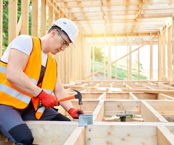 Builder wearing vest an helmet, sitting, holding hammer, beating nail in wooden decking.